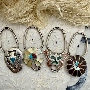 May include: Four silver-toned metal purse hangers with decorative tops labeled 1 through 4. Number 1 is an arrowhead shape with turquoise inlay. Number 2 is a white mother-of-pearl flower. Number 3 is a butterfly with turquoise and coral inlay. Number 4 is a dark brown flower.