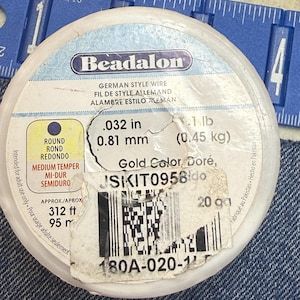 May include: A round spool of Beadalon German style wire in gold color. The label indicates a diameter of .032 inches, 0.81 mm, a length of 312 feet, and a weight of 1 lb. A blue measuring tape is visible at the top showing the spool size.