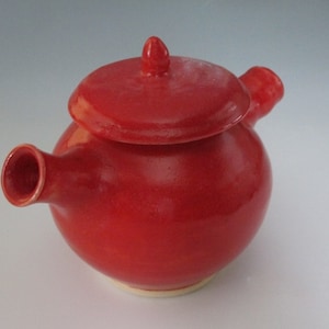 May include: A red ceramic teapot with a lid and a spout. The teapot is glazed and has a smooth finish.