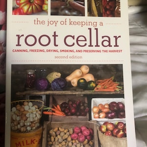 May include: A book titled The Joy of Keeping a Root Cellar, Second Edition, by Jennifer Megyesi with photography by Geoff Hansen. The cover features a collage of fresh produce, canning jars, and wooden crates filled with vegetables and eggs.
