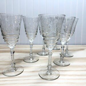 May include: Six clear, etched wine glasses with a floral design. The glasses have a flared bowl, a ribbed stem, and a round base. The glasses are arranged on a light-colored wooden surface.