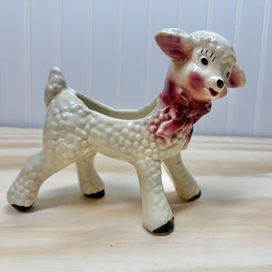 May include: A white ceramic lamb planter with a pink bow. The lamb has a textured surface that resembles wool. The planter has a small opening on the back for drainage.