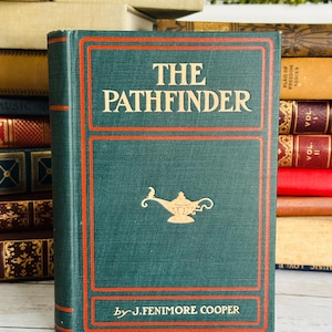 May include: A vintage copy of "The Pathfinder" by J. Fenimore Cooper. The book has a teal cover with red trim and gold lettering. A gold lamp graphic is featured on the cover. The book is part of a collection of antique books.