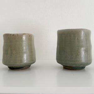 May include: Two ceramic cups with a light green glaze. The cups are slightly different in size and shape.