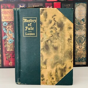 Pode incluir: Um livro antigo intitulado "Mastery of Fate" de Larson. O livro tem uma capa verde-escura com letras douradas e uma seção com padrões dourados. Outros livros antigos são visíveis no fundo.