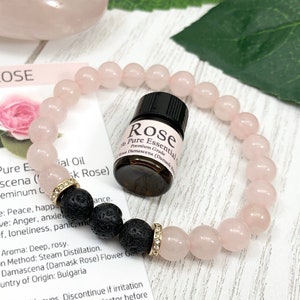 May include: A stretch bracelet featuring smooth, pale pink rose quartz beads, three porous black lava beads, and two gold-toned spacers with small clear crystals. A small amber glass bottle of Rose essential oil sits inside the bracelet loop.