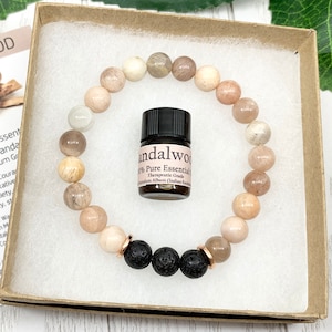 May include: A circular beaded bracelet featuring smooth, light-toned stone beads and three textured black lava beads with rose gold-toned spacers, accompanied by a small amber glass bottle labeled Sandalwood 100% Pure Essential Oil.