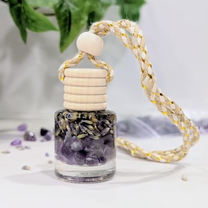 May include: A small clear glass bottle filled with purple amethyst crystals and dried lavender buds. The bottle features a light-colored wooden cap and a braided beige and gold cord for hanging, designed to be used as a car or room air freshener.
