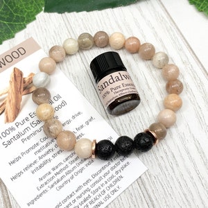 May include: A beaded bracelet featuring smooth, light brown and tan stone beads, three black porous lava beads, and two rose gold-toned metal spacers. A small amber-colored glass bottle labeled Sandalwood 100% Pure Essential Oil sits inside the bracelet loop.