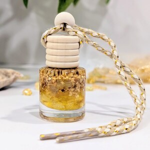 May include: A small glass bottle filled with yellow crystals and small beige fragments, topped with a ribbed light wood cap and a round wooden bead. A braided cord with white, beige, and metallic gold threads is attached for hanging.
