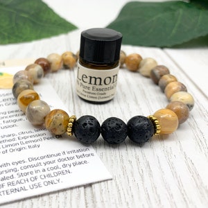 May include: A beaded bracelet featuring tan and brown mottled stone beads, three black porous lava beads, and small gold-toned metal spacers. A small amber glass bottle labeled Lemon 100% Pure Essential Oil sits inside the bracelet loop.