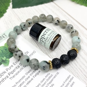 May include: A beaded bracelet featuring light gray speckled stone beads, three black porous lava stone beads, and small gold-toned metal spacers. A small amber glass bottle of peppermint essential oil is placed inside the bracelet loop.