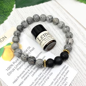 May include: A circular beaded bracelet featuring grey marbled stone beads, two black porous lava stone beads, and small gold-toned metal spacers. A small amber glass bottle labeled Lemon 100% Pure Essential Oil sits inside the bracelet loop.