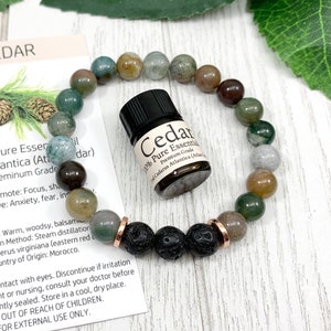 May include: A beaded bracelet featuring round moss agate stones in shades of green, brown, and gray, accented by three black porous lava beads and two rose gold-toned metal spacers. A small amber glass bottle of Cedar essential oil is included.