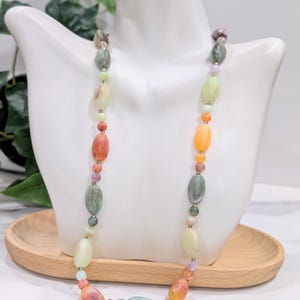 May include: A colorful beaded necklace with oval and round beads in shades of orange, green, yellow, and purple. The necklace is displayed on a white mannequin bust. The beads are strung together with a delicate chain.