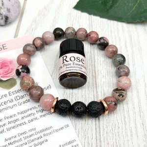 May include: A beaded bracelet featuring round pink and grey rhodonite stones, three black porous lava beads, and two rose gold-toned metal spacers. A small amber glass bottle with a black cap labeled Rose 100% Pure Essential Oil sits in the center.