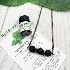 May include: A small bottle of peppermint essential oil next to a necklace with three black lava beads. The necklace has a copper-colored chain. A card with text about the oil is also present.