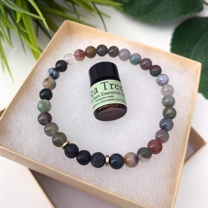 May include: A circular beaded bracelet featuring multicolored smooth stone beads, three porous black lava beads, and two gold-toned metal spacers. A small amber glass bottle of Tea Tree Pure Essential Oil with a black cap sits in the center.