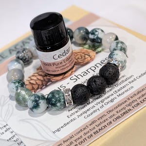 May include: A beaded bracelet with green and white stone beads and black lava beads. A small bottle of Cedar essential oil sits on top of the bracelet. The bottle has a black cap and a white label.