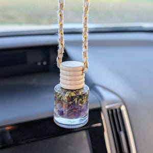 May include: A small, clear glass bottle hanging from a braided beige cord with gold metallic threading. The bottle features a ribbed wooden cap and is filled with a layer of purple amethyst crystals topped with dried green lavender buds.
