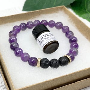 May include: A beaded bracelet featuring smooth purple amethyst spheres, two textured black lava beads, and two gold-toned rhinestone spacers, accompanied by a small brown glass bottle labeled Lavender 100% Pure Essential Oil 2 ml.