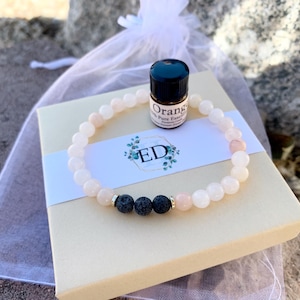 May include: A light pink beaded bracelet with black lava beads and a small bottle of orange essential oil. The bracelet rests on a beige gift box with a white label featuring the initials "ED" and floral accents. A sheer white bag is in the background.
