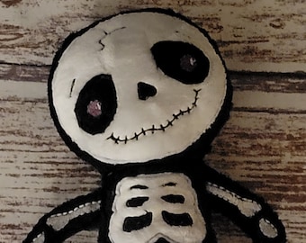May include: A handmade plush doll shaped like a skeleton, crafted from black and white felt. The doll features a round white head with black eye sockets and a stitched mouth, and a black body with white felt bone appliques on the torso and limbs.