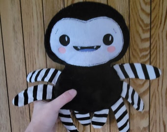 May include: A plush spider toy with a black body and a white face featuring large black eyes, a small blue mouth with two white fangs, and pink cheeks. The spider has eight legs with a black and white horizontal striped pattern throughout.