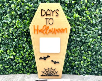 May include: A wooden coffin-shaped Halloween countdown sign with the words "DAYS TO Halloween" in black and orange. Includes a blank white space for writing the number of days, two bat cutouts, and a spiderweb design. A black marker is placed in front.
