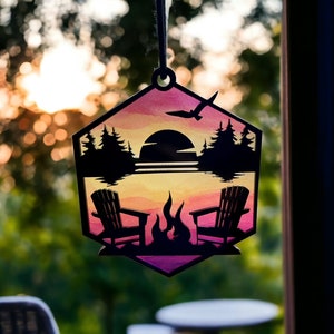 May include: A hexagonal wooden ornament with a sunset scene painted on it. The scene includes a lake, trees, a campfire, and two Adirondack chairs. The ornament is hanging from a string.