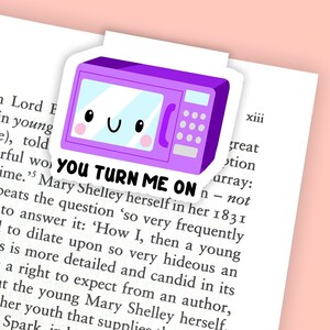 Puede incluir: Un marcapáginas morado con forma de microondas con una cara sonriente y el texto "YOU TURN ME ON". El marcapáginas está en una página de un libro con texto negro. El fondo es rosa claro.