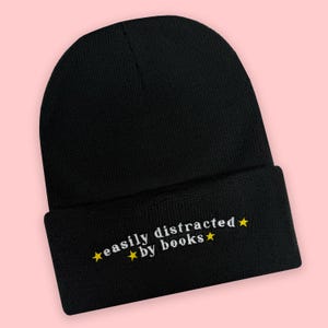 Pode incluir: Um gorro de malha preto com dobra. A dobra apresenta um texto bordado em branco que diz "easily distracted by books" com pequenas estrelas amarelas colocadas entre as palavras e nas extremidades da frase, criando um design gráfico simples.