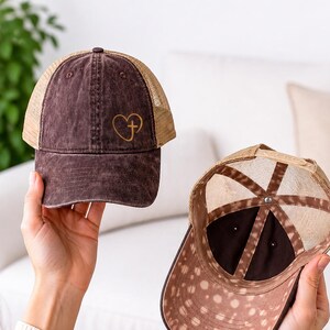 May include: Two brown baseball caps with mesh backs are displayed. One cap has a gold heart and cross design. The other cap has a spotted pattern on the underside of the brim. The caps are held up by a person.