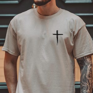 May include: A beige t-shirt with a black cross graphic on the left chest. The cross is a simple, stylized design with a slightly distressed look.