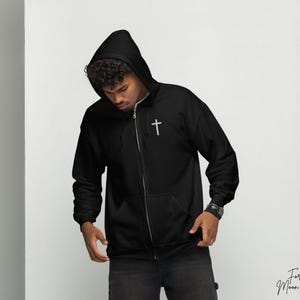 May include: A black zippered hoodie with a small silver cross on the chest. The hoodie has a hood and a zipper closure. The model is wearing the hoodie and looking down.