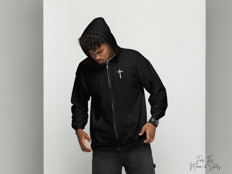 May include: A black zippered hoodie with a small silver cross on the chest. The hoodie has a hood and a zipper closure. The model is wearing the hoodie and looking down.