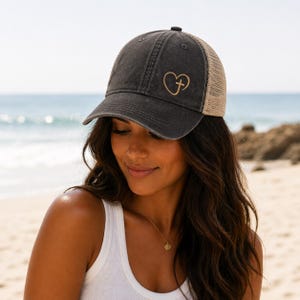 May include: A charcoal gray trucker hat with a tan mesh back. The hat features a gold heart and cross design on the front. The hat is worn by a person on a beach.