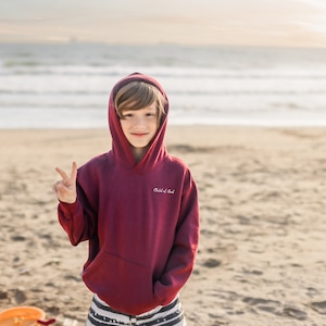 Puede incluir: Una persona joven con una sudadera con capucha color granate con el texto "Child of God" en la parte delantera, de pie en una playa de arena con el oc&eacute;ano de fondo. Sonr&iacute;e y hace el signo de la paz con la mano derecha.