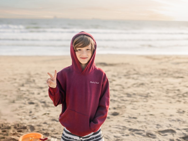Puede incluir: Una persona joven con una sudadera con capucha color granate con el texto "Child of God" en la parte delantera, de pie en una playa de arena con el oc&eacute;ano de fondo. Sonr&iacute;e y hace el signo de la paz con la mano derecha.