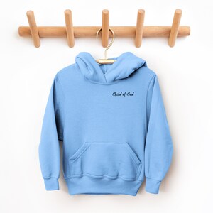 Puede incluir: Una sudadera con capucha azul claro con un bolsillo y una frase bordada en negro "Child of God" en la parte delantera.