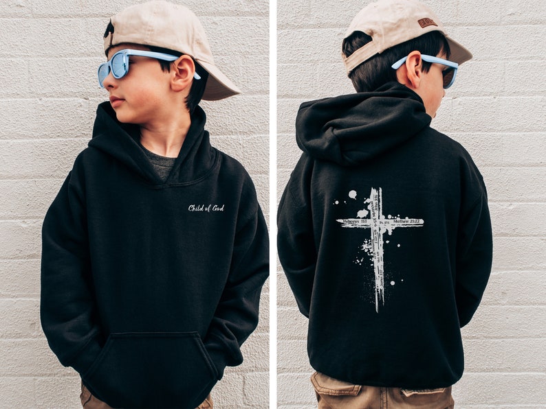 Puede incluir: Sudadera con capucha negra con un dise&ntilde;o de cruz blanca en la espalda y el texto "Child of God" en la parte delantera. La sudadera est&aacute; siendo modelada por un ni&ntilde;o que lleva una gorra de b&eacute;isbol beige y gafas de sol azules.