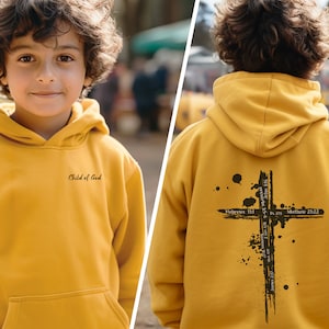 Puede incluir: Una sudadera con capucha amarilla con un gr&aacute;fico de cruz negra en la espalda y el texto "Child of God" en la parte delantera.