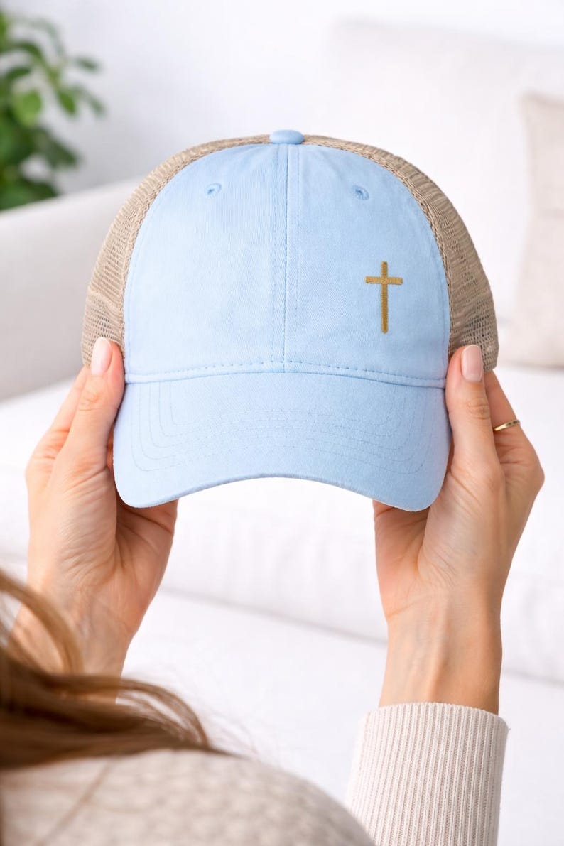 Pu&ograve; includere: Cappellino da camionista azzurro con retro in rete beige e visiera curva. Il cappellino presenta una croce dorata ricamata sul davanti. Il cappellino &egrave; tenuto in mano da una persona.