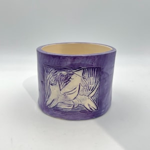 May include: A cylindrical ceramic pot in a vibrant purple hue. The exterior features a carved design of a figure and starfish. The interior is a light beige colour. This handmade pot is suitable for plants or as a decorative item.