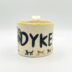May include: Hand-painted ceramic mug with the word "DYKE" in bold black letters. The mug features a yellow rim and a small handle. Illustrations of dogs are at the bottom of the mug. A person is illustrated on the side of the mug.