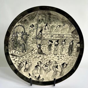 Puede incluir: Plato de cerámica en blanco y negro con una ilustración pintada a mano de una reunión de figuras con sombreros puntiagudos. La escena representa una figura diabólica y un grupo de personas en una mesa, con otras figuras bailando abajo.