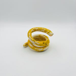 May include: A coiled ceramic snake sculpture. The snake is primarily yellow with white and light green accents. The snake's head has a pink mouth and small black eyes. The sculpture is on a white background.