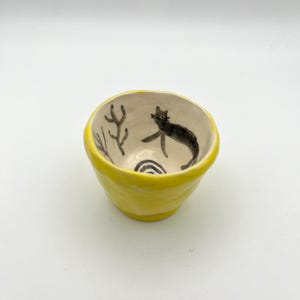 以下が含まれることがあります： 鮮やかな黄色の外側とクリーム色の内側を持つ、手描きの小さなセラミックボウル。内側には、黒猫の気まぐれなイラストと、抽象的な植物とスパイラルのデザインが描かれています。このボウルは、小さなアイテムを入れるのに最適です。