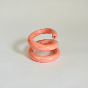 May include: A quirky, handmade ceramic sculpture in a spiral form. The sculpture is a soft coral colour with a smiling face on the top. The piece is a unique decorative item.