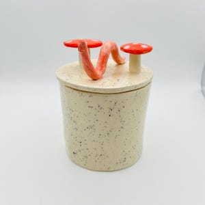 以下が含まれることがあります： 斑点のあるベージュ色のセラミックジャー。蓋には、赤いキノコ2つとサンゴ色のミミズが飾られています。ジャーは円筒形で、小さなアイテムの保管に最適です。